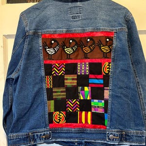 May include: A blue denim jacket with a colorful patchwork design on the back. The patchwork features a brown panel with bird illustrations and a grid of various patterned squares in red, black, and other colors.