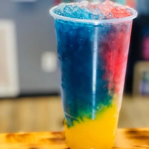 Puede incluir: Un vaso alto de pl&aacute;stico transparente lleno de una bebida colorida en capas. La bebida presenta capas vibrantes de amarillo, verde, azul y rojo, cubiertas con hielo picado. El vaso est&aacute; sobre una superficie de madera.