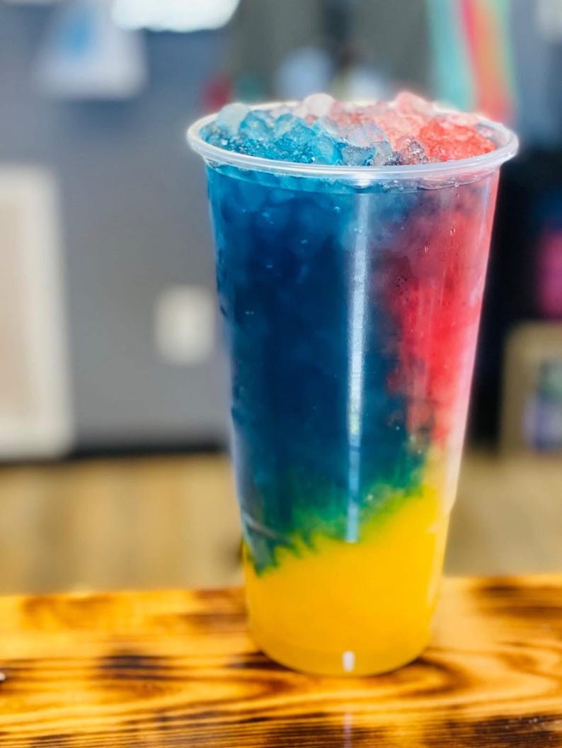 Puede incluir: Un vaso alto de pl&aacute;stico transparente lleno de una bebida colorida en capas. La bebida presenta capas vibrantes de amarillo, verde, azul y rojo, cubiertas con hielo picado. El vaso est&aacute; sobre una superficie de madera.