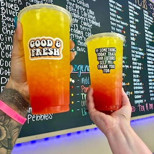 Puede incluir: Dos vasos de pl&aacute;stico transparente llenos de una bebida en capas amarilla, naranja y roja. El vaso m&aacute;s grande tiene una pegatina "GOOD & FRESH", y el vaso m&aacute;s peque&ntilde;o tiene una pegatina que dice "DO SOMETHING TODAY THAT YOUR FUTURE SELF WILL THANK YOU FOR".