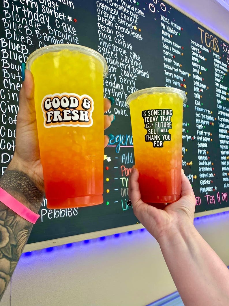 Puede incluir: Dos vasos de pl&aacute;stico transparente llenos de una bebida en capas amarilla, naranja y roja. El vaso m&aacute;s grande tiene una pegatina "GOOD & FRESH", y el vaso m&aacute;s peque&ntilde;o tiene una pegatina que dice "DO SOMETHING TODAY THAT YOUR FUTURE SELF WILL THANK YOU FOR".