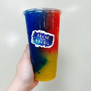 Puede incluir: Una bebida colorida en un vaso de pl&aacute;stico transparente, con capas de azul, rojo, verde y amarillo. Una pegatina con el texto "PROVE THEM WRONG" est&aacute; adherida al vaso. El vaso es sostenido por una mano.