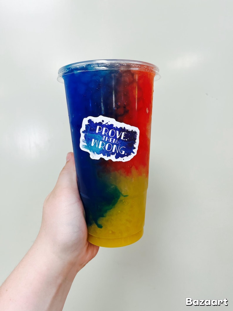 Puede incluir: Una bebida colorida en un vaso de pl&aacute;stico transparente, con capas de azul, rojo, verde y amarillo. Una pegatina con el texto "PROVE THEM WRONG" est&aacute; adherida al vaso. El vaso es sostenido por una mano.