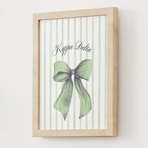 Puede incluir: Impresión enmarcada con una ilustración de acuarela de un lazo verde con cintas fluidas. El fondo tiene rayas verticales en verde claro y blanco. Las palabras "Kappa Delta" están escritas en una elegante escritura.