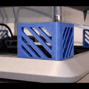 Peut inclure: Gros plan sur une structure de support bleue imprimée en 3D avec un motif en treillis diagonal. Le support est en forme de cube et est positionné sur une surface gris clair. L'image montre une partie d'une imprimante 3D.