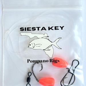 May include: Clear plastic bag containing fishing tackle. The bag features the text "SIESTA KEY" and "Pompano Rigs" with a fish graphic. The tackle includes hooks, swivels, and bright orange floats.