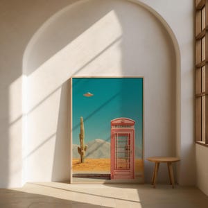 May include: Framed art print featuring a desert landscape with a red telephone booth, a cactus, and a flying saucer in the sky. The artwork has a surreal, retro vibe with a light wooden frame and a small wooden side table.