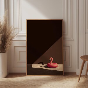 May include: A framed print featuring a red inflatable flamingo pool float. The flamingo is positioned on a light-colored surface against a dark brown background. The print is in a wooden frame, and the scene is lit by a diagonal beam of light.
