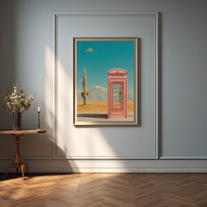May include: A framed art print depicts a pink telephone booth in a desert landscape with a cactus and a UFO. The artwork is displayed on a light gray wall, above a small wooden table with a vase of flowers and a candle.