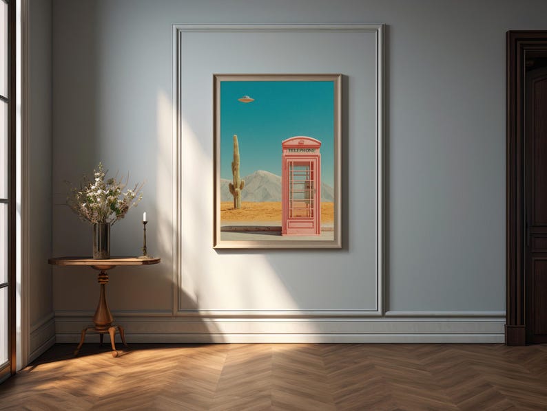 May include: A framed art print depicts a pink telephone booth in a desert landscape with a cactus and a UFO. The artwork is displayed on a light gray wall, above a small wooden table with a vase of flowers and a candle.
