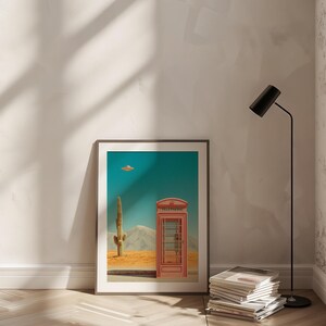 May include: Framed art print featuring a desert landscape with a red telephone booth, a cactus, and a flying saucer against a teal sky. The print is displayed on a white wall, with a black floor lamp and a stack of magazines nearby.