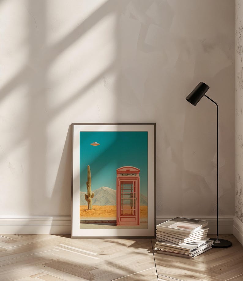 May include: Framed art print featuring a desert landscape with a red telephone booth, a cactus, and a flying saucer against a teal sky. The print is displayed on a white wall, with a black floor lamp and a stack of magazines nearby.