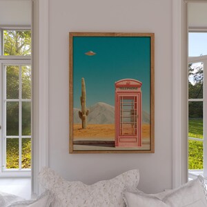 May include: A framed art print featuring a surreal desert landscape. A pink telephone booth stands in the desert with a cactus and mountains in the background. A flying saucer hovers in the turquoise sky. The print is displayed in a room with windows.