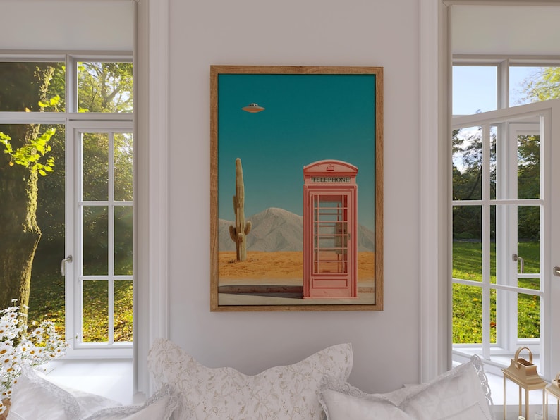 May include: A framed art print featuring a surreal desert landscape. A pink telephone booth stands in the desert with a cactus and mountains in the background. A flying saucer hovers in the turquoise sky. The print is displayed in a room with windows.