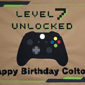 May include: A brown banner with black and green accents, featuring the text "LEVEL 7 UNLOCKED" and "Happy Birthday Colton!". A black game controller is in the center. The design has a video game theme.