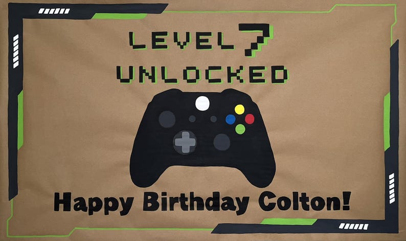 May include: A brown banner with black and green accents, featuring the text "LEVEL 7 UNLOCKED" and "Happy Birthday Colton!". A black game controller is in the center. The design has a video game theme.