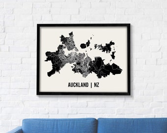 May include: Black and white map print of Auckland, New Zealand, with the city name and country abbreviation in the bottom right corner. The map is framed in a black frame and is hanging on a white brick wall.
