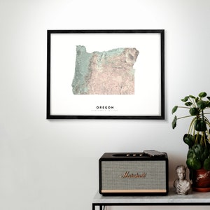 May include: A framed print of a topographic map of Oregon in muted green, pink, and brown colours. The map is surrounded by a black frame and the word "OREGON" is printed in black below the map.