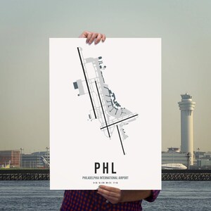 May include: A white poster featuring a black and grey airport layout map of Philadelphia International Airport (PHL). The poster displays the airport code "PHL" and the airport name "Philadelphia International Airport" in black text.