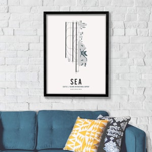K&ouml;nnte beinhalten: Ein schwarzer gerahmter Druck mit wei&szlig;em Hintergrund und einem grauen und blauen Flughafenlayout. Der Text "SEA" ist in gro&szlig;en, fetten, blauen Buchstaben. Unter dem Flughafenlayout steht der Text "Seattle | Tacoma International Airport".