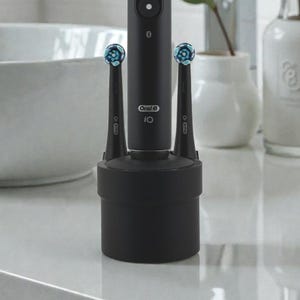 Könnte beinhalten: Eine schwarze elektrische Zahnbürste mit zwei Bürstenköpfen in einer Ladestation. Die Zahnbürste hat einen Ein-/Ausschalter und der Markenname "Oral-B iO" ist sichtbar. Die Bürstenköpfe sind blau und weiß. Die Umgebung ist ein Badezimmer.