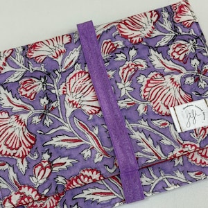 Puede incluir: Estuche de tela con estampado floral en morado, rojo y blanco, asegurado con una banda elástica morada. El diseño presenta flores y hojas estilizadas repetidas. Se adjunta una pequeña etiqueta blanca con texto manuscrito.