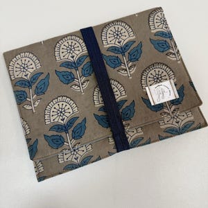 Puede incluir: Funda rectangular de tela con un estampado floral en tonos topo, blanco y azul. El diseño presenta flores y hojas estilizadas. Una banda elástica azul marino asegura la funda y una pequeña etiqueta blanca está adjunta.