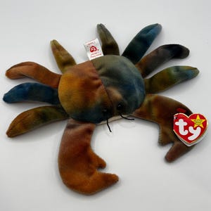 Puede incluir: Un juguete de peluche con forma de cangrejo con un estampado tie-dye colorido en tonos marrones, azules y verdes. El juguete tiene múltiples patas y un par de ojos negros pequeños. Una etiqueta roja y blanca en forma de corazón está adjunta.