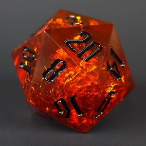 Op de afbeelding: Een twintigzijdige dobbelsteen van doorschijnende oranje hars met glinsterende goudkleurige insluitingen aan de binnenkant. De dobbelsteen heeft scherpe geometrische randen en diepzwarte gegraveerde cijfers, met het getal twintig duidelijk zichtbaar.