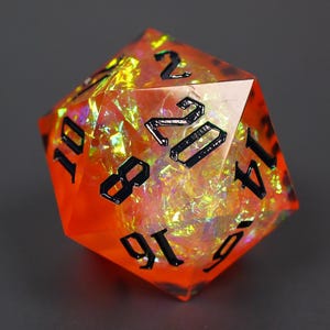 Pode incluir: Um dado de vinte lados feito de resina laranja translúcida com flocos de folha iridescente embutidos que captam a luz. O dado apresenta bordas afiadas e números gravados em preto profundo, com o número vinte exibido na face frontal.