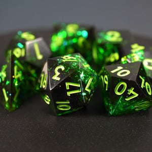 Könnte beinhalten: Ein Set aus polyedrischen Würfeln mit einer durchscheinenden smaragdgrünen Harzbasis, in die schimmernde grüne Glitzerpartikel eingebettet sind. Jeder Würfel hat scharfe Kanten und ist mit hellen lindgrünen Zahlen graviert.