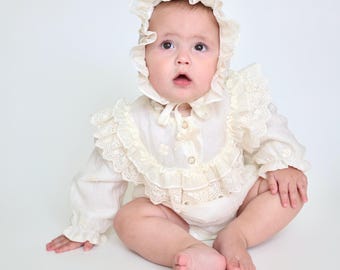 May include: An off-white long-sleeved romper featuring a ruffled collar with intricate eyelet lace trim and matching buttons. The outfit includes a coordinating ruffled bonnet with a tie closure, crafted from a lightweight fabric.
