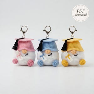 May include: Three crochet graduation gnome keychains. Each gnome wears a different colored graduation cap: pink, blue, and yellow. They have black tassels, white bodies, and hold a diploma. The keychains are attached to silver metal clasps.
