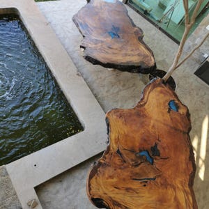 Op de afbeelding: Twee houten tafels met een natuurlijke rand, met een natuurlijke houtnerf. De tafels hebben blauwe harsaccenten en staan op een betonnen terras. Een kleine boom groeit ertussen, met een waterpartij in de buurt.