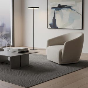 Può includere: Una poltrona imbottita color beige con schienale curvo, un tavolino da caffè grigio a profilo basso con una pila di libri e una lampada da terra nera, alta e sottile, con testa circolare, su un tappeto grigio scuro testurizzato.