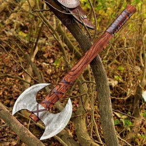 Könnte beinhalten: Eine Doppelklingen-Axt mit Holzgriff und lederumwickeltem Griff. Die Axtköpfe sind silberfarben, und der Griff hat ein Holzmaserungsmuster. Die Axt wird im Freien gezeigt, die an einem Ast lehnt.