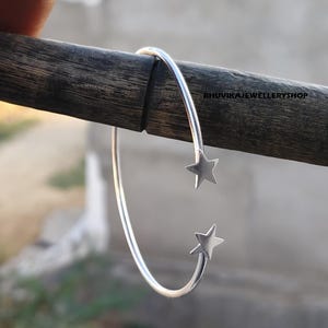 Peut inclure: Un bracelet ouvert argenté orné d'étoiles. Ce bracelet présente un design minimaliste, avec une bande incurvée et une étoile à chaque extrémité. Le bracelet est présenté sur un fond flou.