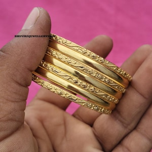 May include: A set of six gold bangles with floral and leaf patterns. The bangles are held in a hand against a pink background. The jewelry is shiny and appears to be made of metal.