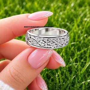 Könnte beinhalten: Ein silberfarbener Metallring mit einem komplizierten, geprägten Blumenmuster, bei dem detaillierte Blätter und Blüten das gesamte Band umschließen. Der Text BHUVIKAJEWELLERYSHOP ist in kleinen schwarzen Buchstaben über dem Ring gedruckt.