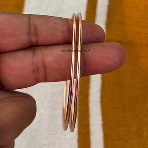 May include: Two delicate rose gold bangles. The thin, circular bracelets are displayed against a yellow and orange background. The jewelry is simple and elegant, suitable for everyday wear. The shop name "BHUVIKAJEWELRYSHOP" is visible.