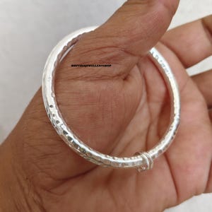 May include: A silver bangle bracelet with a hammered texture. The bracelet is round and has a simple clasp. The text "BHUVIKAJEWELLERYSHOP" is visible on the bracelet. The bangle is held in a hand.