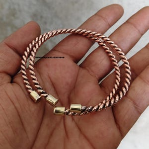 May include: Two copper-colored, twisted rope-style bracelets with gold-colored clasps. The bracelets are held in a hand. The text "BHUVINJJEWELLERYSHOP" is visible in the background.