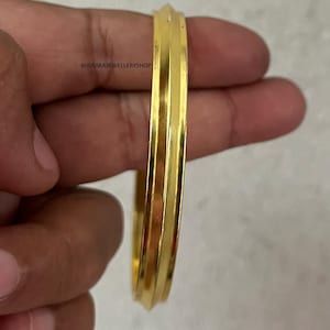 Puede incluir: Un brazalete dorado con un diseño acanalado. La joya se sostiene en una mano, mostrando su superficie pulida y su estilo simple y elegante. El brazalete es una pieza de joyería clásica.