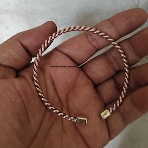 May include: A copper-colored twisted metal bracelet with gold-colored end caps. The bracelet is open and appears to be unworn. The jewelry is held in a hand. The shop name "BHUVIKAJEWELLERYSHOP" is visible.