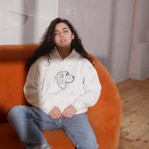 May include: A white hooded sweatshirt featuring a black line art illustration of a dog's head on the chest, paired with light blue denim jeans and white trainers. The hoodie has long sleeves, a front pocket, and a drawstring hood.
