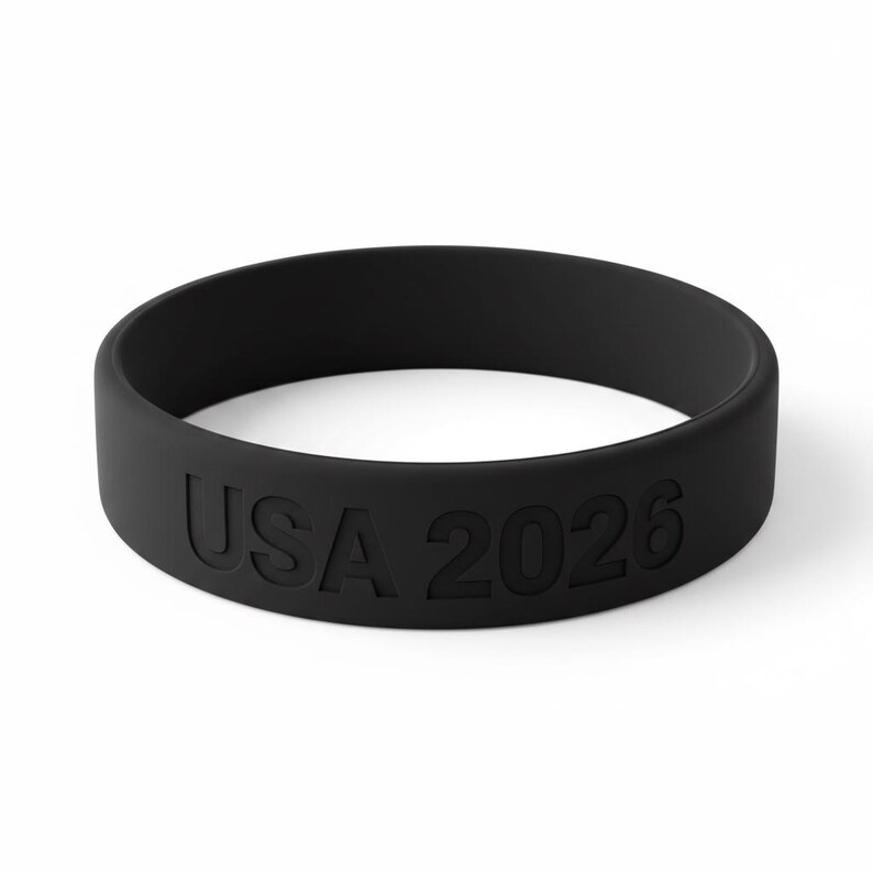 May include: A black rubber wristband with the embossed text "USA 2026". The band is a solid, matte black color and appears to be made of a flexible material. The text is in a sans-serif font.