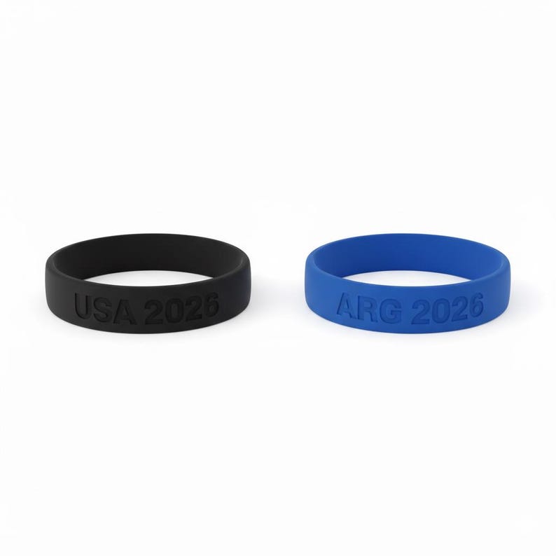 May include: Two silicone wristbands, one black and one blue. The black band has "USA 2026" embossed on it, while the blue band reads "ARG 2026". Both bands are circular and have a smooth texture.
