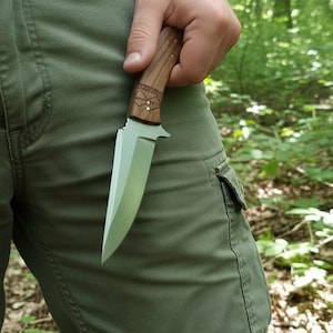 May include: A hunting knife with a wooden handle and a silver blade. The handle has decorative carvings and brass accents. The knife is held against a background of green pants and a forest.