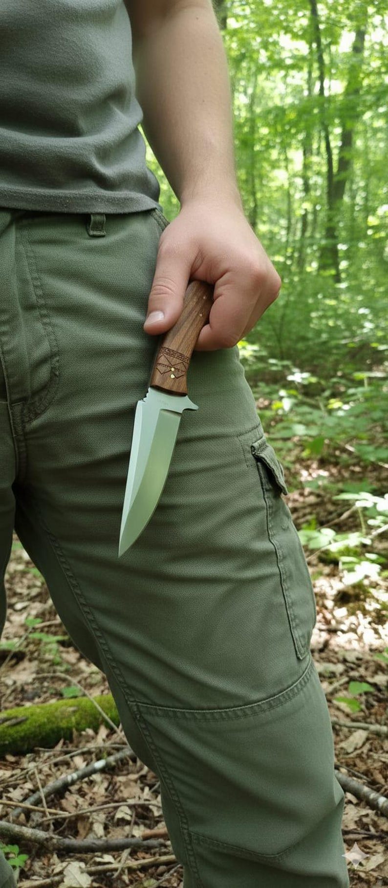 May include: A hunting knife with a wooden handle and a silver blade. The handle has decorative carvings and brass accents. The knife is held against a background of green pants and a forest.