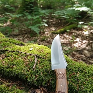 May include: A handcrafted knife with a polished blade and a wooden handle featuring decorative etchings. The knife rests on a moss-covered log in a forest setting, showcasing its outdoor utility and craftsmanship.
